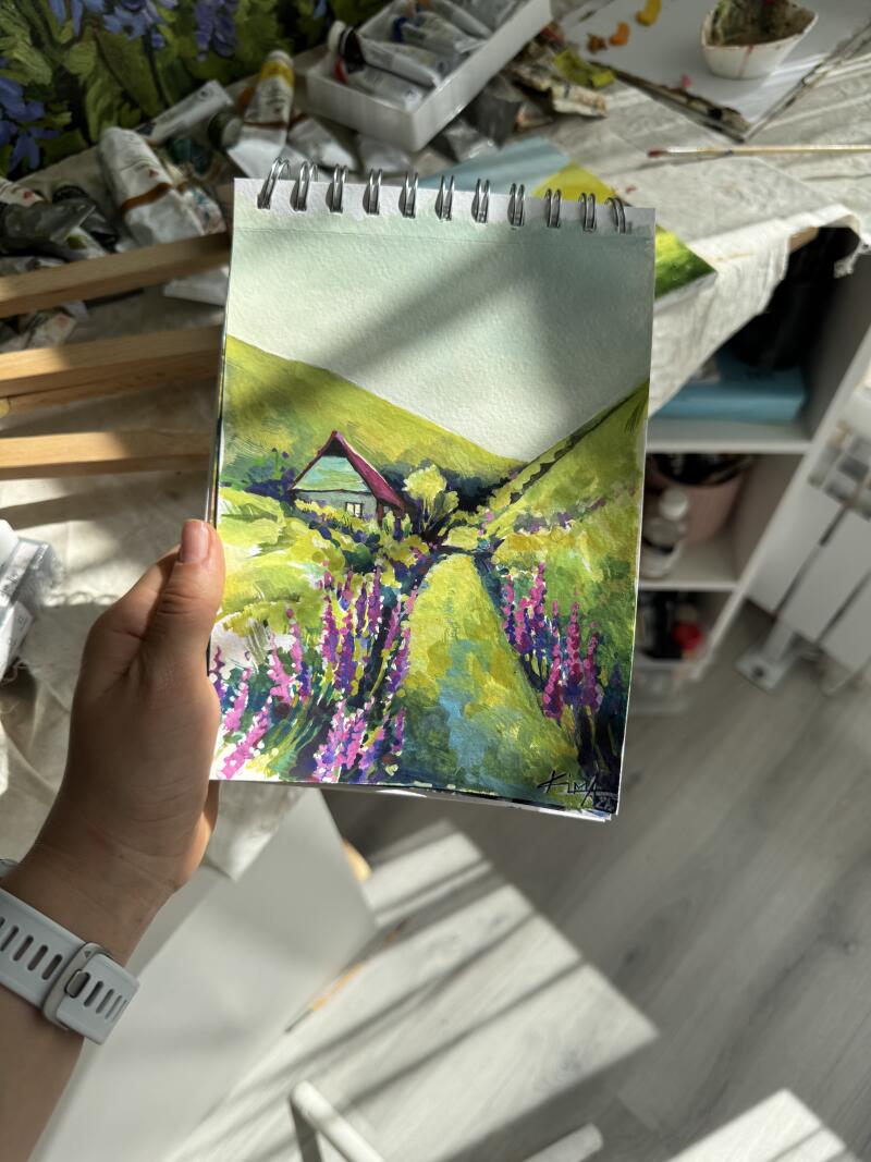 Рука держит альбом на спирали с акварельной картиной яркого пейзажа с зелеными холмами, небольшим домиком с красной крышей и тропинкой, усаженной фиолетовыми цветами.