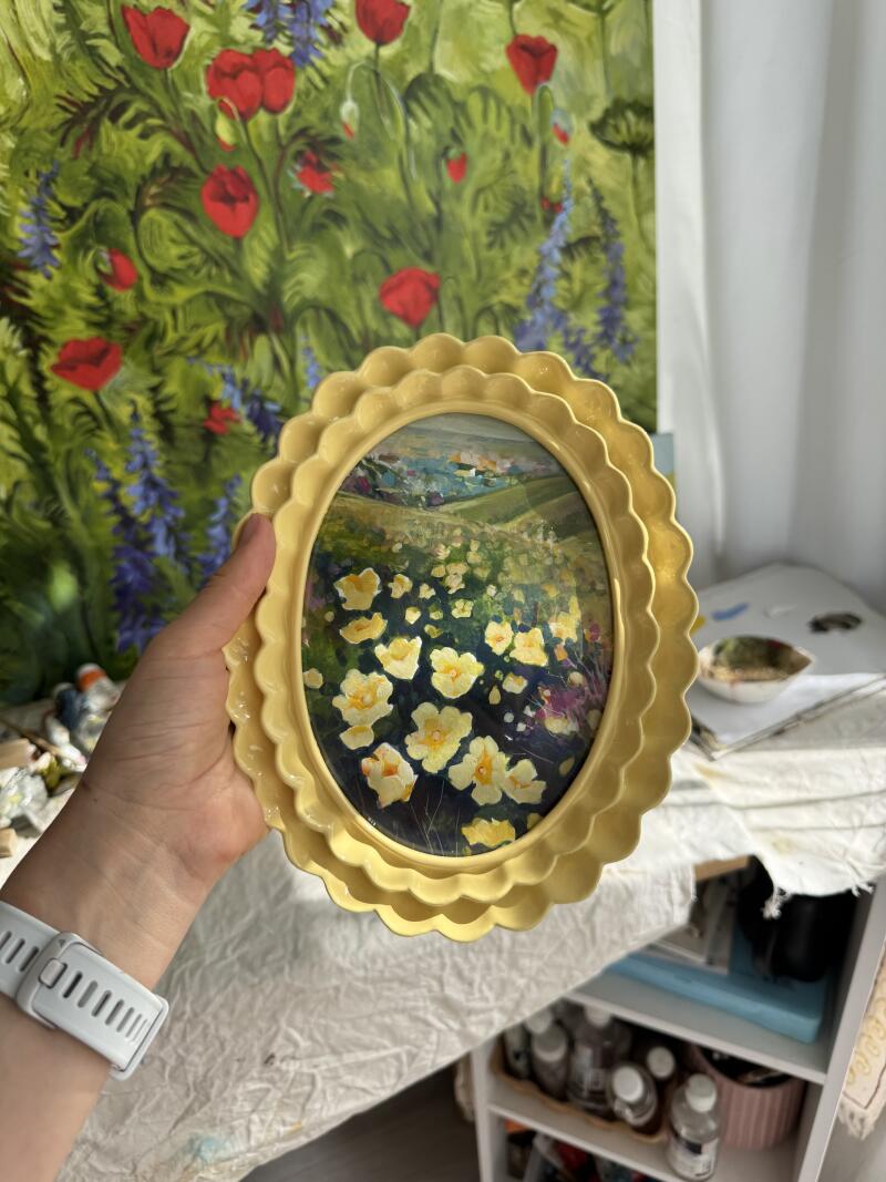 Рука держит картину в рамке с изображением желтых цветов в поле с пейзажем на заднем плане; на заднем плане большая картина с красными цветами и синими растениями.