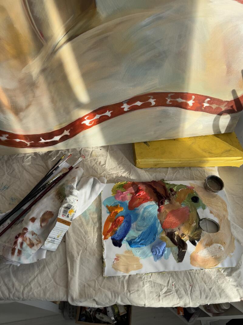 На фотографии изображено рабочее место художника, включающее частично завершенную картину с красной и белой декоративной каймой, палитру с различными цветами краски, несколько кистей, тюбик краски и желтую коробку, все это расположено на текстурированной белой поверхности.