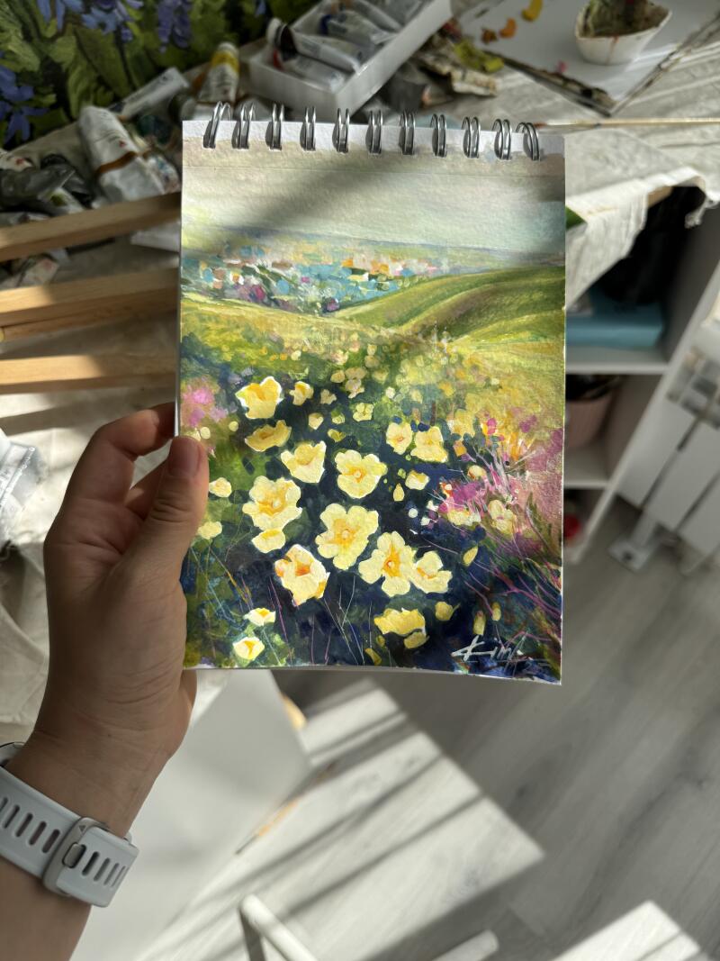 Рука держит альбом для рисования с акварельной картиной пейзажа, на котором изображены желтые цветы на переднем плане, зеленый холм на среднем плане и городской пейзаж на заднем плане.