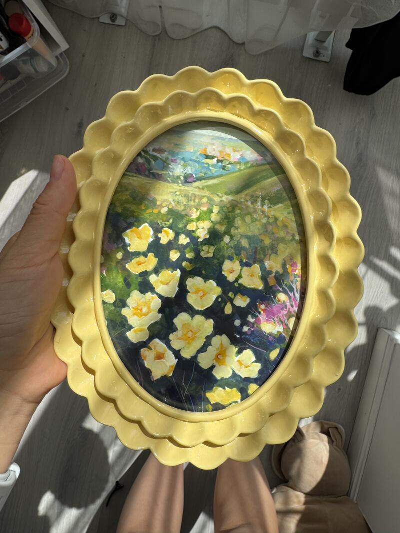 Человек держит овальную картину в желтой рамке с фестонами, изображающую пейзаж с желтыми цветами на переднем плане.