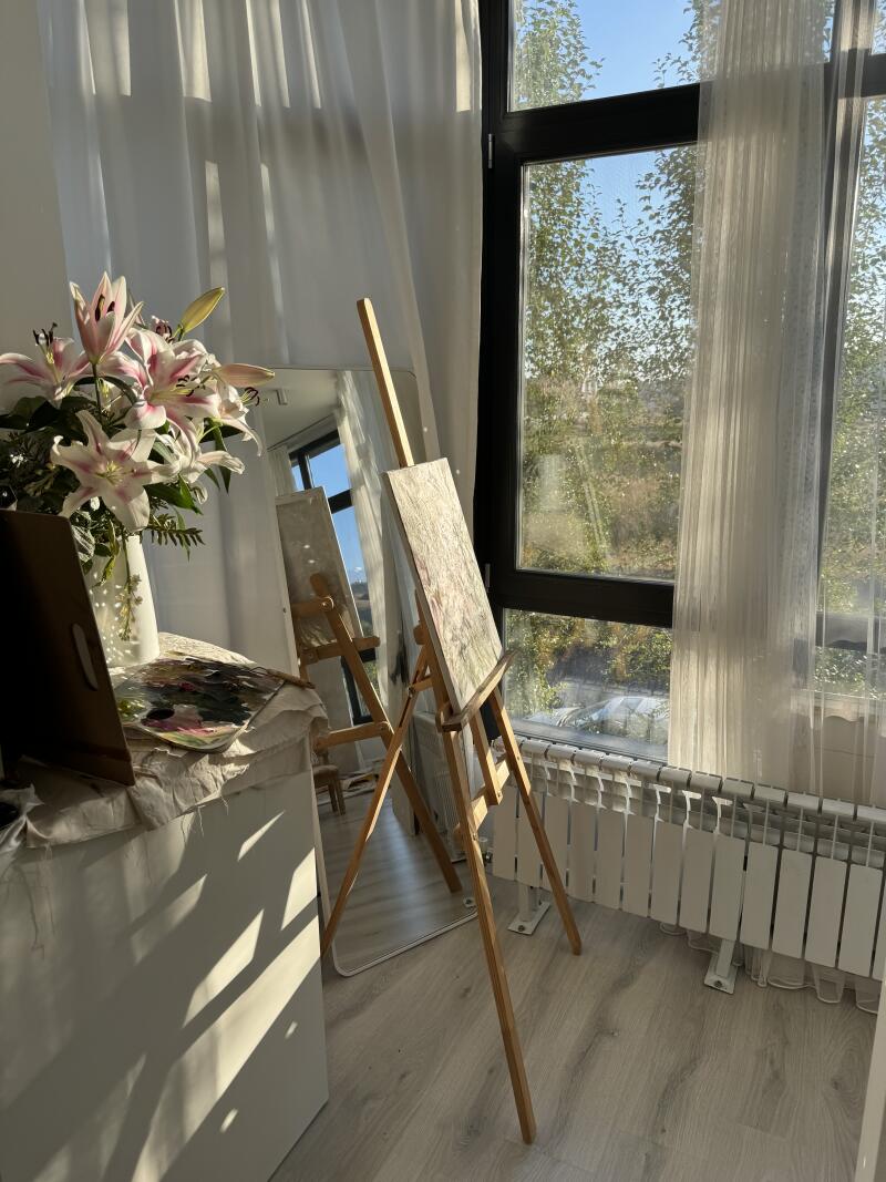 На снимке изображена художественная студия с двумя мольбертами, на одном из которых картина, ваза с лилиями и палитра, все залито солнечным светом возле окна с прозрачными белыми шторами.
