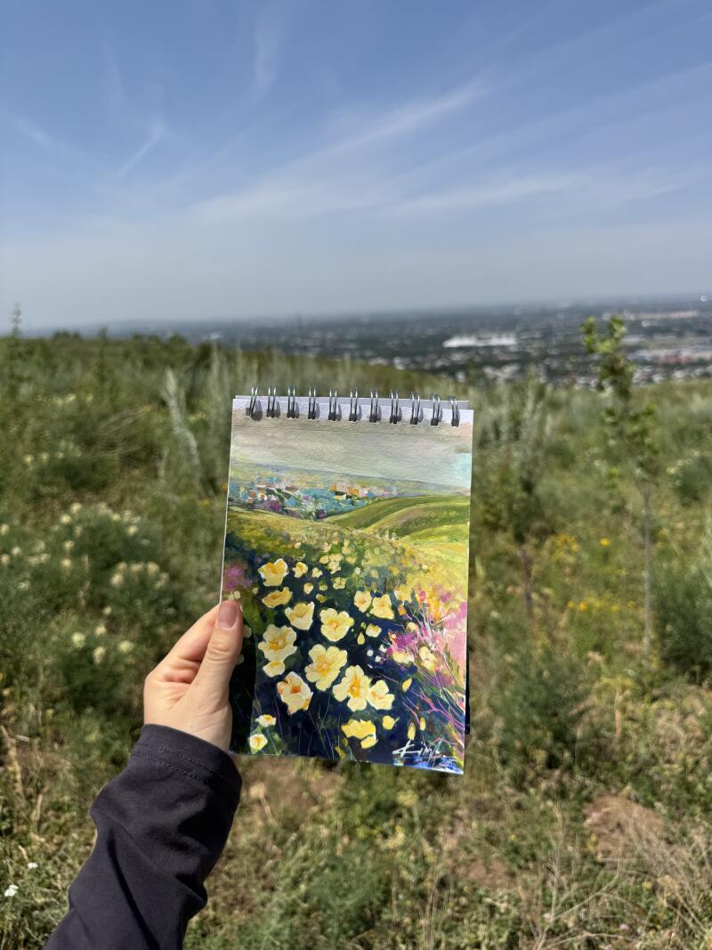 Рука держит альбом для рисования на спирали, на котором изображена красочная картина поля желтых цветов и отдаленного городского пейзажа на фоне травянистого склона холма под голубым небом.