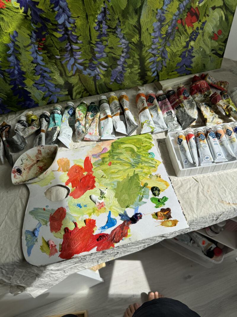 На изображении палитра со смешанными масляными красками, множество тюбиков с краской и картина с цветами на заднем плане.