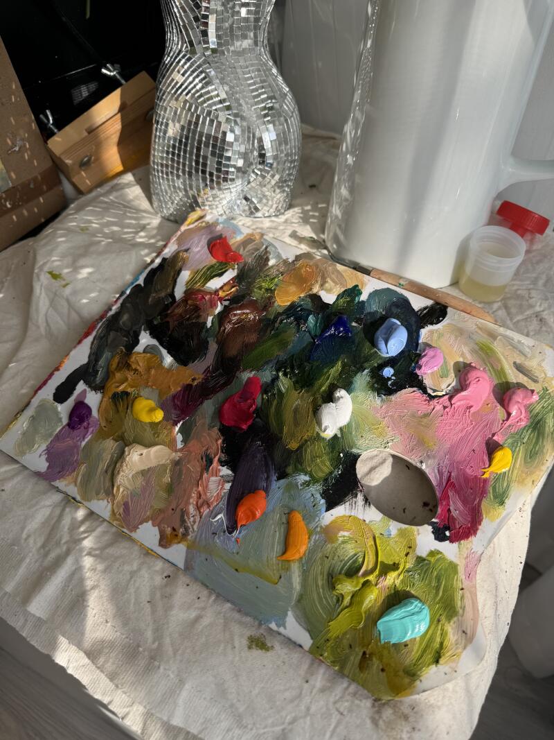 Палитра художника с различными цветами масляной краски стоит рядом с зеркальной вазой и белым контейнером.