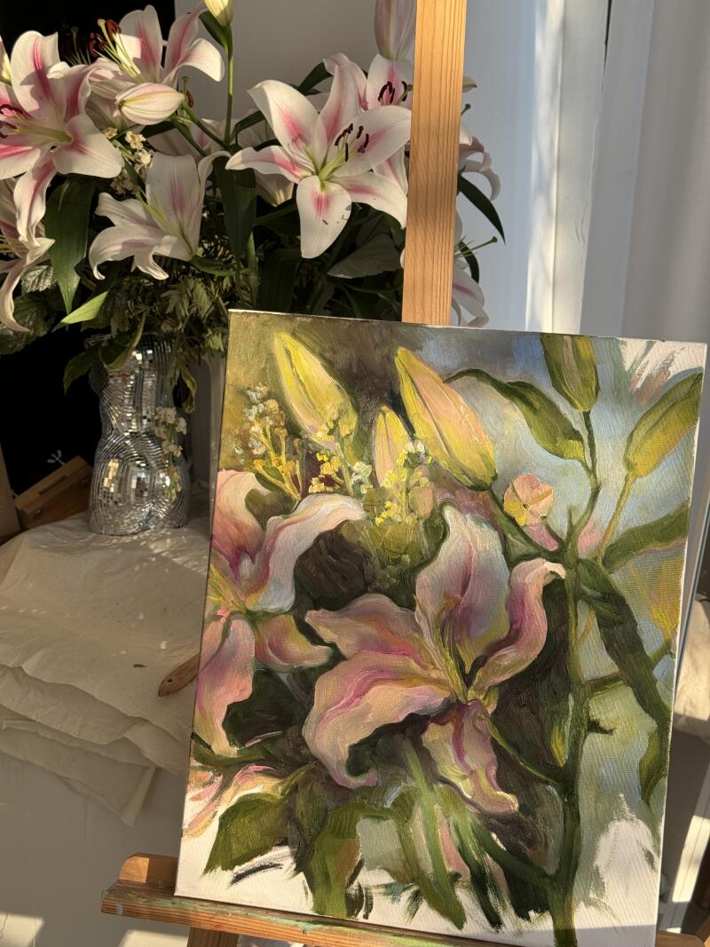 На изображении представлена картина розовых и белых лилий на мольберте, с вазой аналогичных лилий на заднем плане.