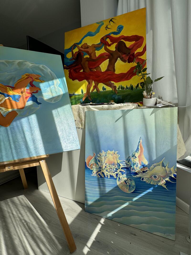 На изображении представлены три картины: одна изображает человеческую фигуру в синих тонах, другая - танцующие фигуры в красных и синих шарфах на желто-зеленом фоне, и третья - морские ракушки на синем фоне, все выставлены в помещении.