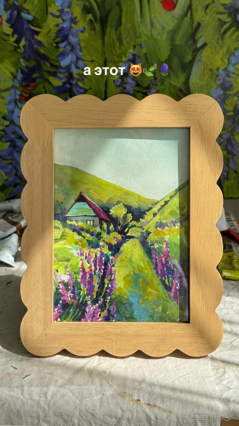 На картине в рамке изображен пейзаж с домом, зелеными холмами и фиолетовыми цветами.