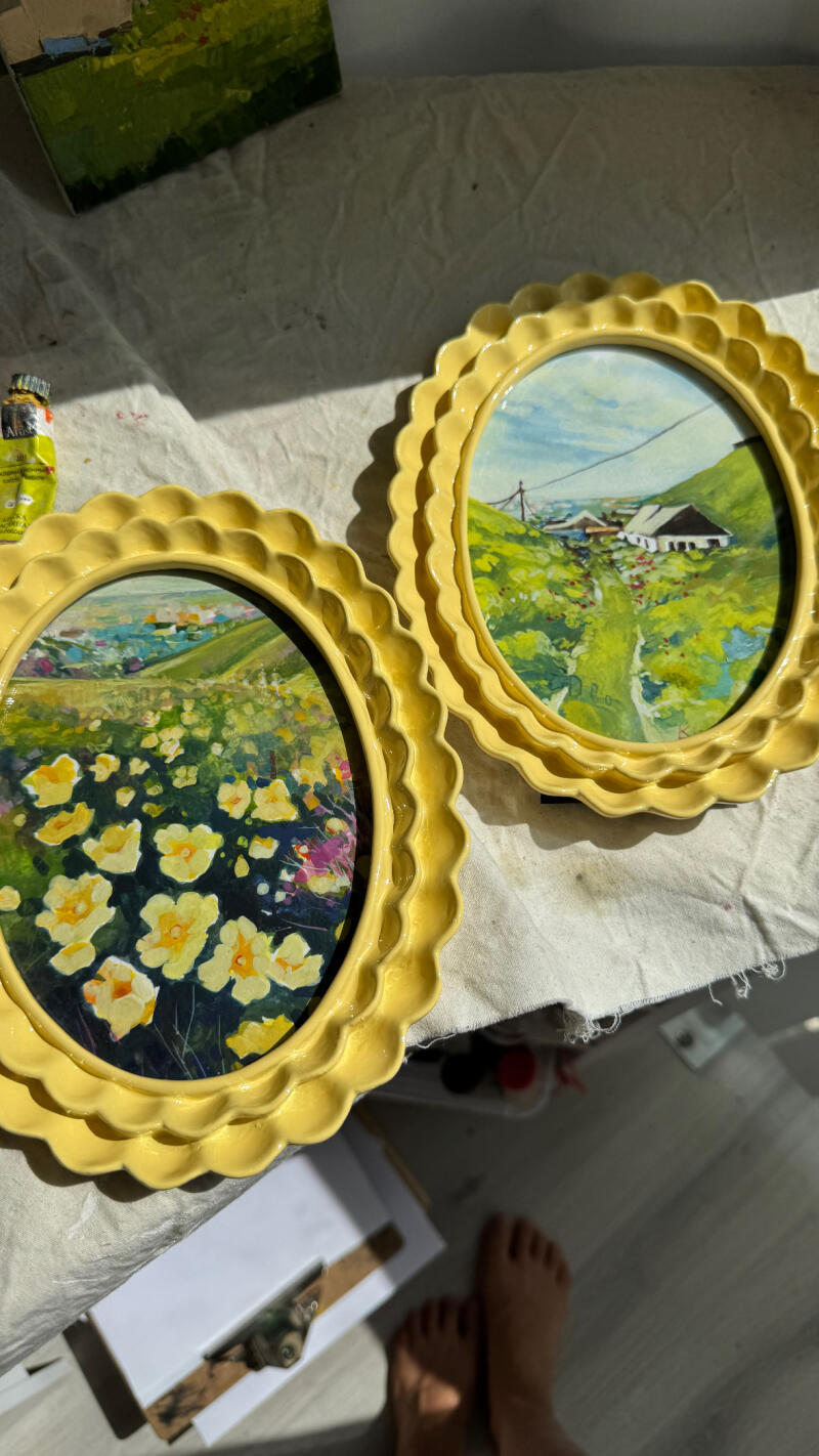 На изображении представлены две обрамленные пейзажные картины с желтыми зубчатыми рамками на белой поверхности, одна из которых изображает поле желтых цветов, а другая - зеленые холмы со зданиями.