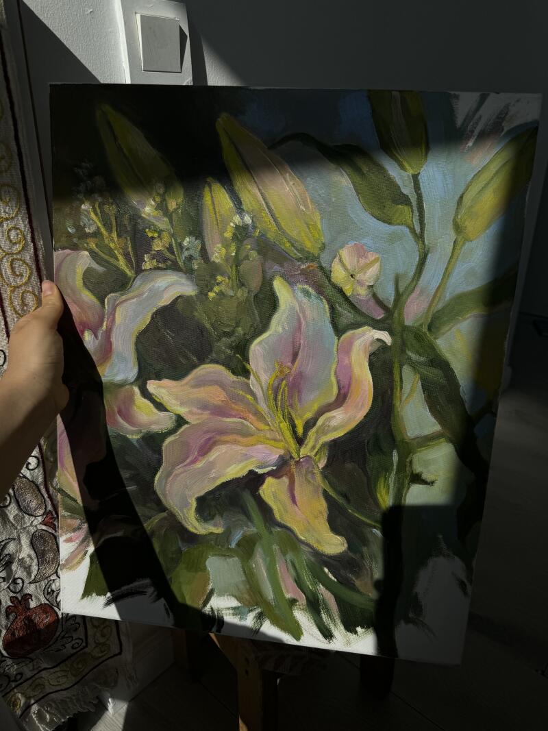 Рука держит картину с изображением лилий в светлой комнате, демонстрируя белые и желтые цветы на фоне синего и зеленого.