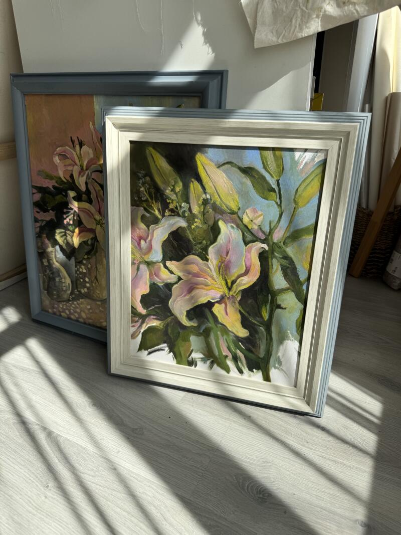 Две картины в рамках прислонены к стене, обе изображают цветочные композиции с лилиями.