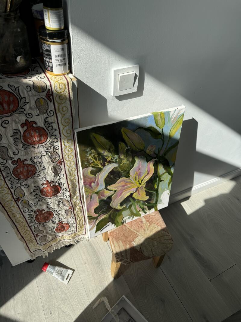 На изображении картина с лилиями, прислоненная к маленькому табурету, рядом со столом, покрытым узорчатой тканью, вокруг разбросаны художественные принадлежности, все залито солнечным светом.