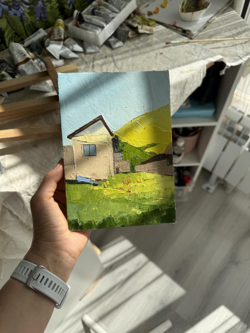 Человек держит небольшую картину с изображением дома на фоне желтого холма и голубого неба, выполненную толстыми мазками.
