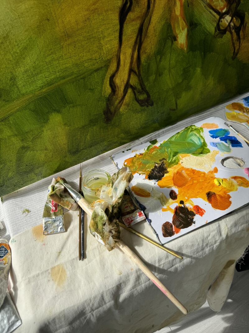 Крупный план показывает картину в процессе работы с палитрой зеленых, желтых, оранжевых и коричневых красок, а также кистями и банкой с прозрачной жидкостью, лежащими на запачканной краской ткани.