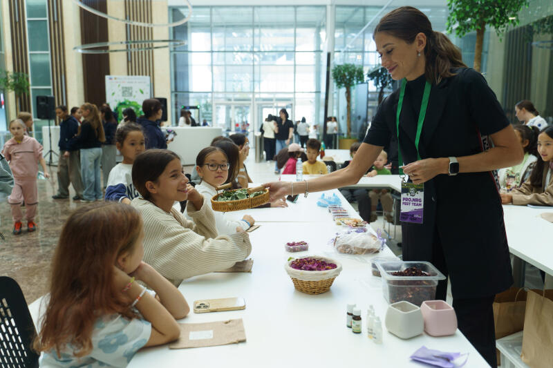 Женщина с бейджем «PRO.NRG HOST TEAM» раздает корзину с зелеными листьями группе детей, сидящих за столом, заполненным различными материалами, включая сушеные цветы и маленькие бутылочки.