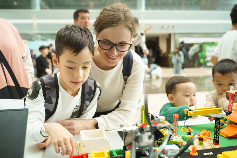 Женщина в очках и маленький мальчик увлеченно играют с кубиками Lego, рядом с другими детьми.