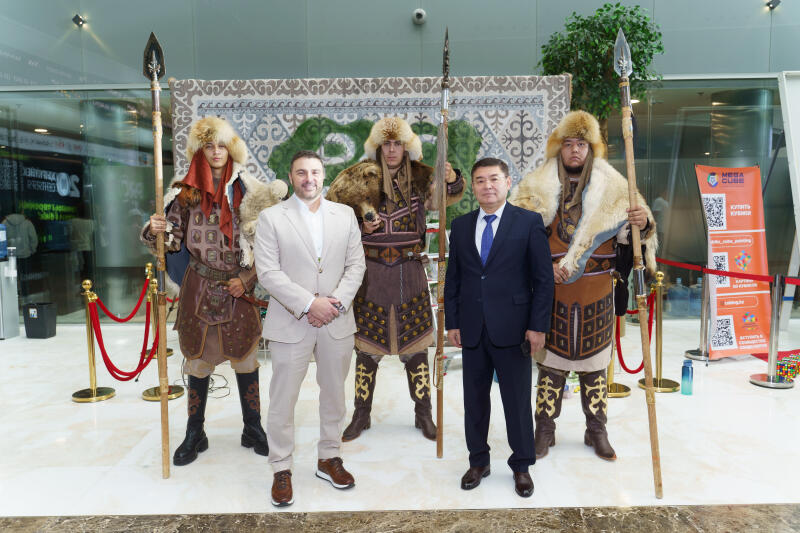 На фотографии изображены двое мужчин в костюмах, стоящие перед тремя людьми, одетыми в традиционные казахские воинские костюмы, держащими копья, на фоне узорчатого ковра.