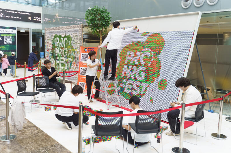 Группа людей собирает большую мозаику из кубиков Рубика, чтобы сформировать текст "PRC NRG FEST". Один человек стоит на лестнице и добавляет кубики в верхнюю часть мозаики, в то время как другие сидят или стоят на коленях, работая над отдельными кубиками или секциями.