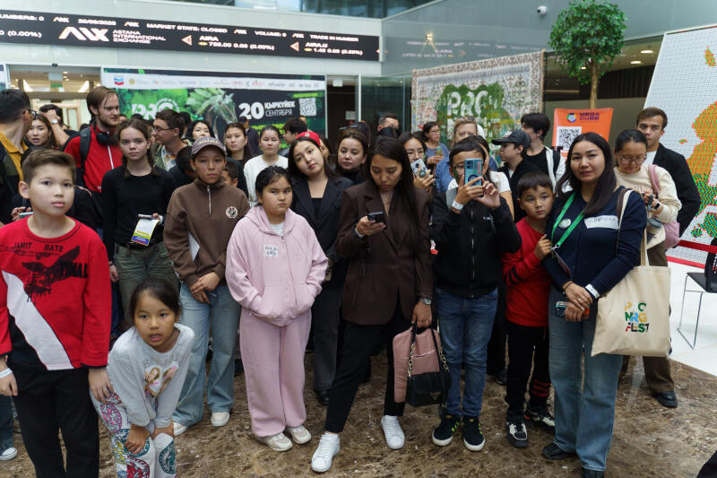 Группа людей, включая взрослых и детей, собралась в помещении возле тикера фондового рынка, отображающего финансовые данные; некоторые смотрят в телефоны или фотографируют. Обстановка включает элементы публичного мероприятия или собрания, на заднем плане видны вывески и декоративные объекты. На вывеске виден текст «20 Қыркүйек Сентября».