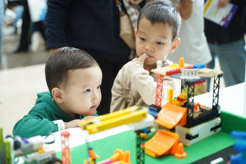 Два ребенка играют с разноцветными строительными блоками, один держит палец во рту, сосредоточившись на создании.