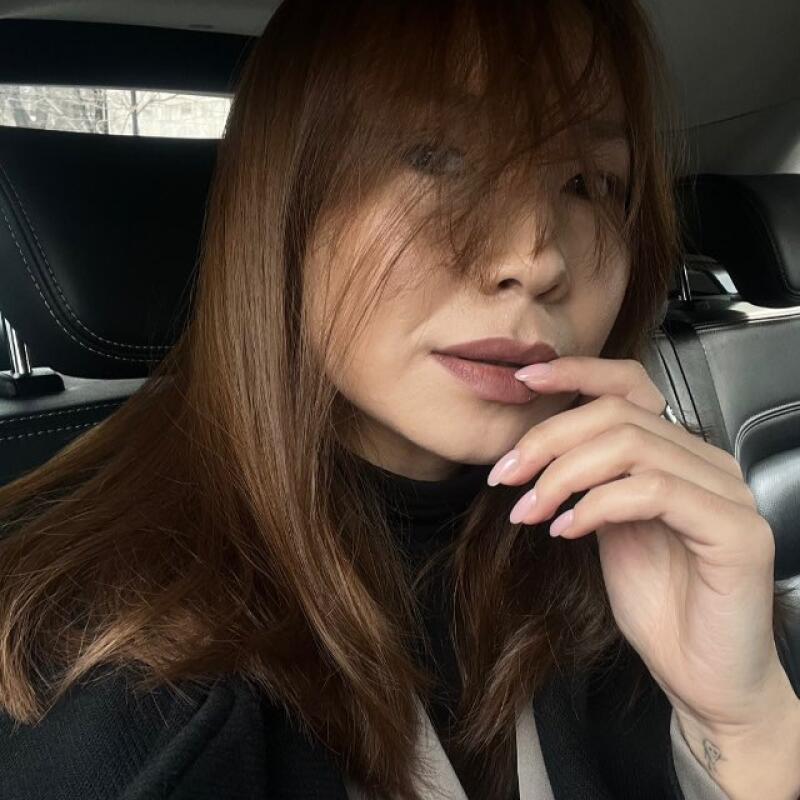 A woman with long brown hair and a black turtleneck is sitting in a car with a seatbelt, holding her finger to her lips.