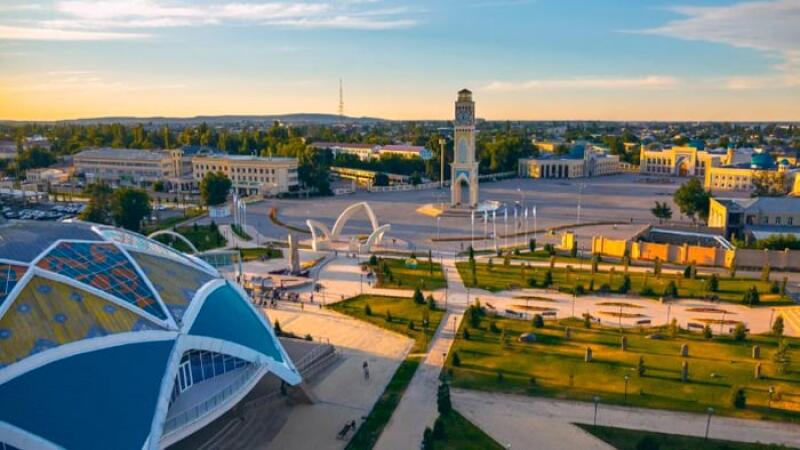 This is a scenic view of a city square featuring a large clock tower, green spaces, and various architectural structures under a partly cloudy sky.
