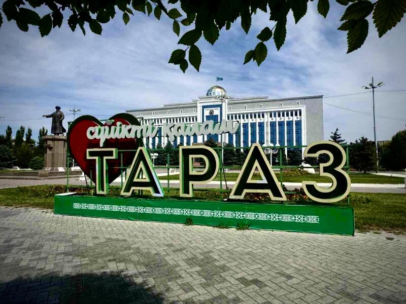 A sign that reads "Cүйікті қалам Тараз" and "ТАРАЗ" is in a park in front of a building.