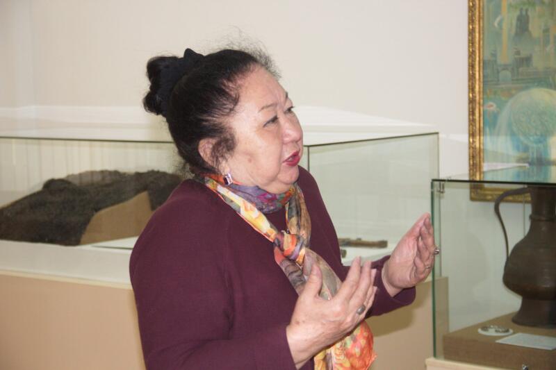 An older woman with dark hair pulled back is speaking, wearing a maroon sweater and a colorful scarf, standing in front of glass display cases, one containing a dark, lumpy object and another featuring a metal jug.