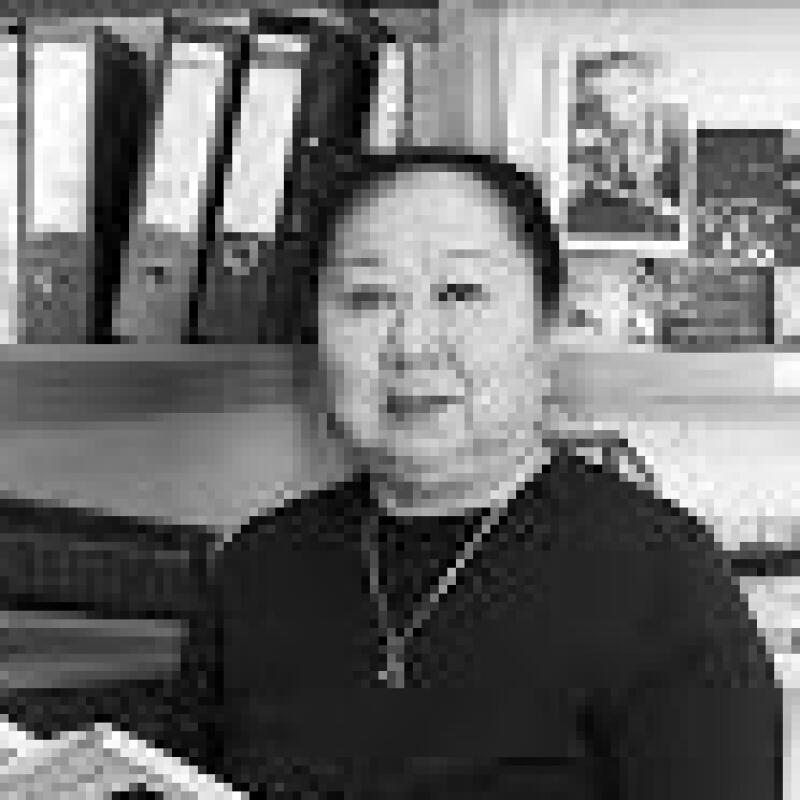 A grayscale portrait of an Asian woman wearing a dark shirt and a necklace with a pendant. Behind her are binders and photos on a shelf.