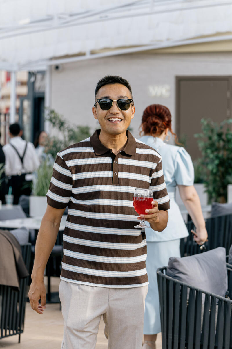 A smiling man in sunglasses and a brown and white striped polo shirt holds a red drink in an outdoor setting, with other people and a 'WC' sign visible in the background.