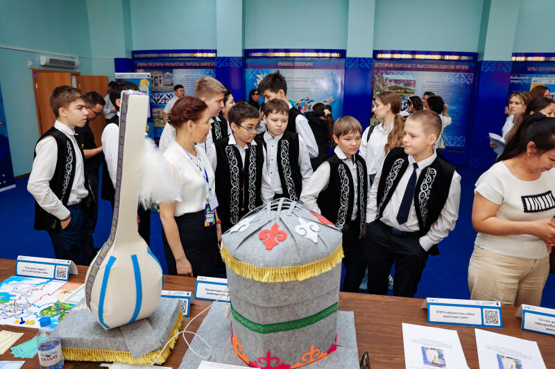 The image shows a group of students and adults gathered around a table displaying handmade models of Kazakh cultural symbols, including a dombra and a yurt.