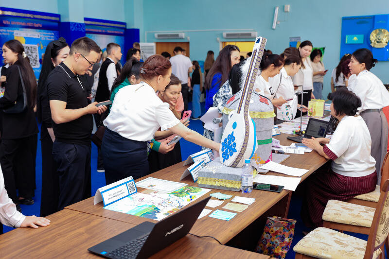 A group of people are gathered around a table at an event, looking at items such as a dombra model, a map, and QR codes; a laptop is also on the table.