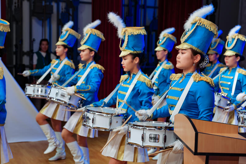 A group of young women are dressed in bright blue marching band uniforms and hats with white plumes while playing white drums.