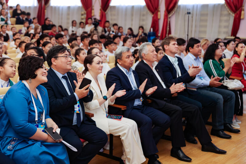 A group of people, including children, are seated and clapping in an auditorium with red curtains.