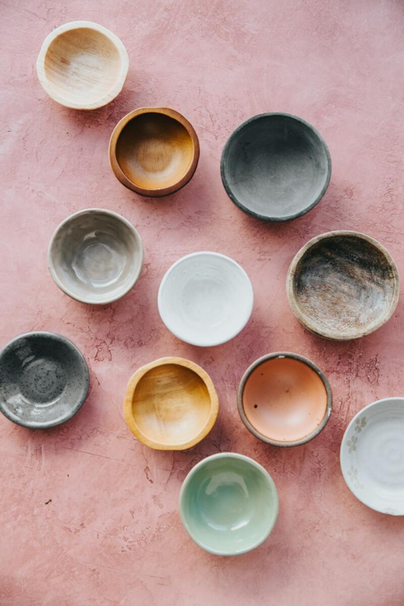 A top-down view of eleven small, empty bowls made from various materials and colors, including wood, ceramic, and clay, scattered across a textured pink surface.