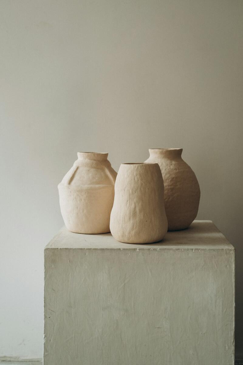 Three light beige, textured ceramic vases of varying shapes stand on a light-colored concrete block against a plain light wall.