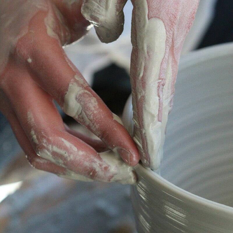 Close-up of hands covered in light-colored clay, shaping a white or light gray pottery piece on a wheel.
