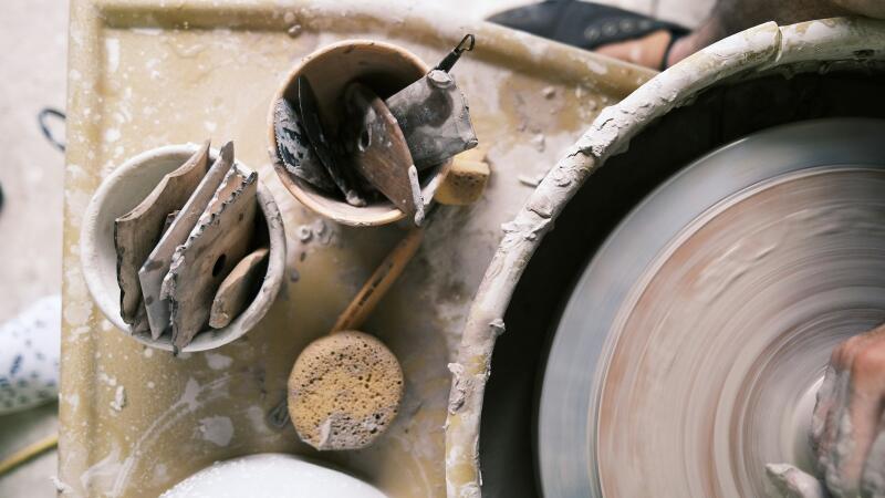 An overhead view of a pottery wheel with a person's hands shaping clay, surrounded by various pottery tools like sponges and scrapers on a wet, clay-splattered surface.