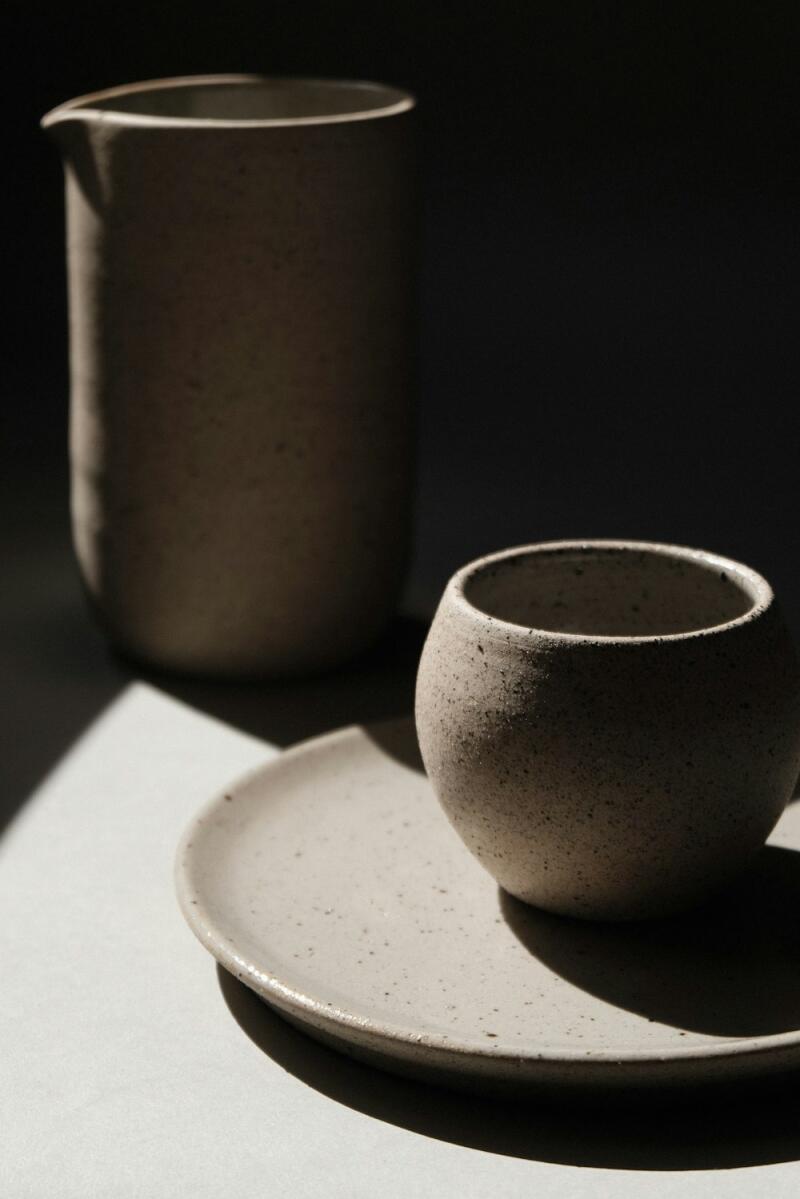 A tall, light-colored ceramic pitcher stands on the left, next to a smaller, round ceramic cup on a saucer, all with a speckled texture, illuminated by strong directional light creating sharp shadows.