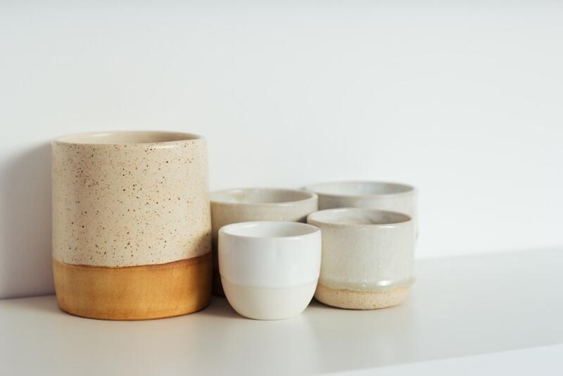 A collection of handmade ceramic cups and a tall vase-like object in neutral tones, including speckled beige, light brown, white, and off-white, arranged on a white surface against a white background.
