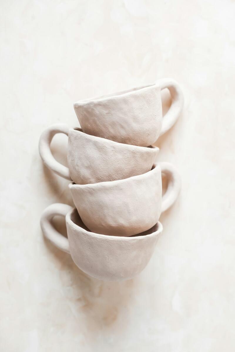 Four light beige, handmade ceramic cups are stacked vertically on a light neutral background. The cups have a rustic, textured appearance.
