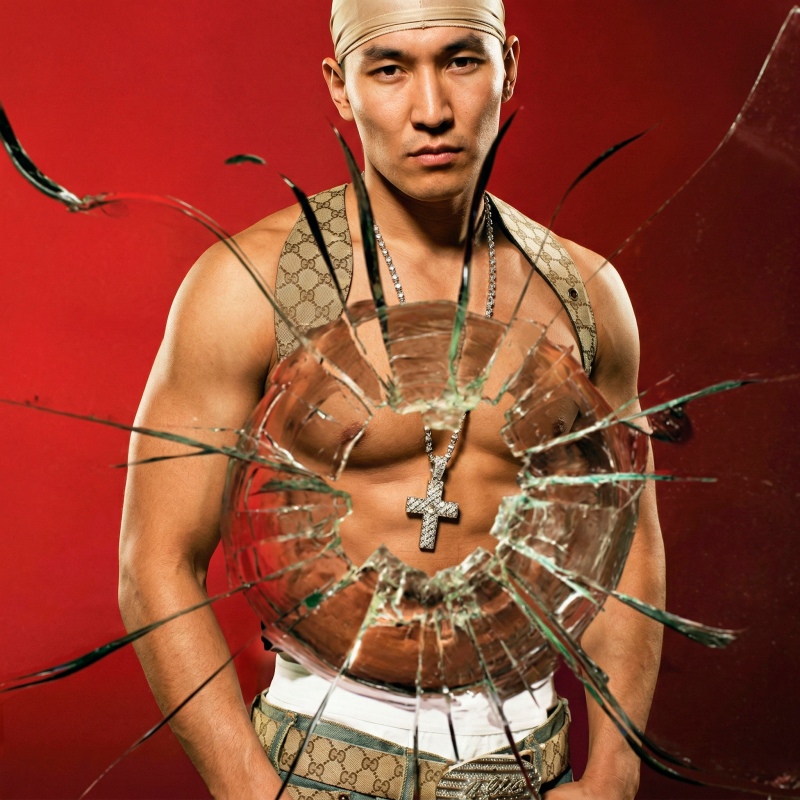 An Asian man with a gold headscarf, chain, and cross necklace stands in front of a red background behind a shattered glass pane.