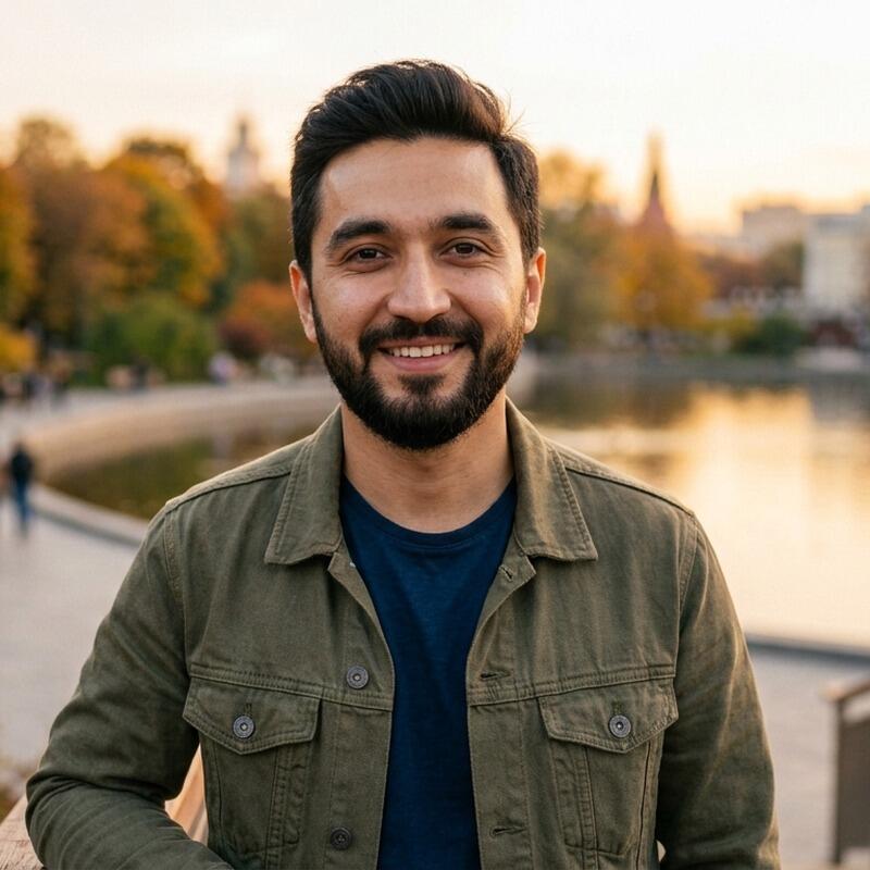 A smiling man with dark hair and a beard wears a green jacket over a blue t-shirt, standing outdoors with a blurred background of autumn trees, water, and buildings under a golden sky.