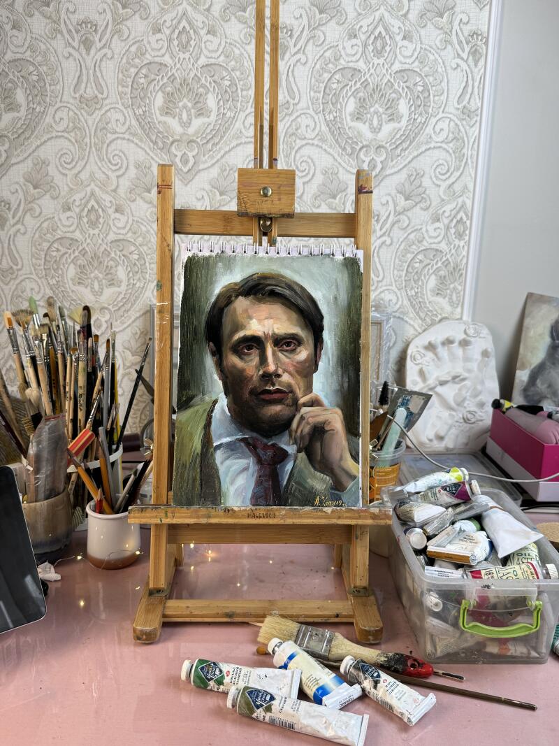 The image shows a painting of Mads Mikkelsen on a wooden easel, with paint tubes and brushes scattered around it. The painting is signed "A. Kozlov" and "HALEVICI".