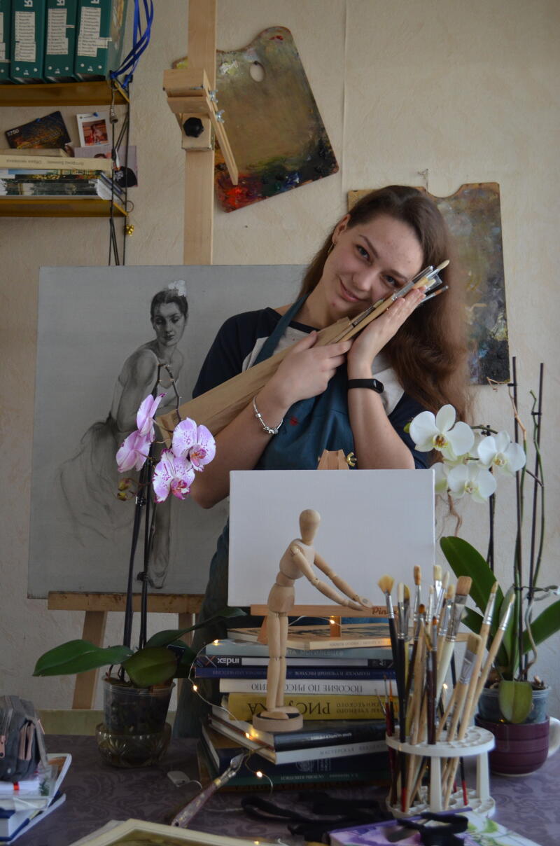 A woman in a blue apron is smiling and holding paintbrushes near her face in front of a painting of a woman, a wooden mannequin on books, and potted orchids.