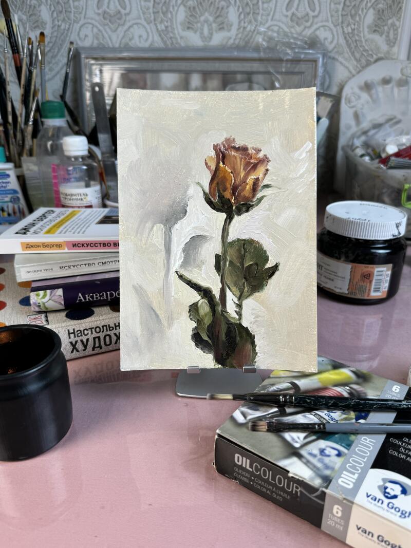 The image features a painting of a rose on a canvas, with visible brushstrokes creating a textured effect, set against a backdrop of art supplies and books, including texts in Russian, such as "ИСКУССТВО" (Art) and "Акварель" (Watercolor); a box of "van Gogh OILCOLOUR" paints is also visible.