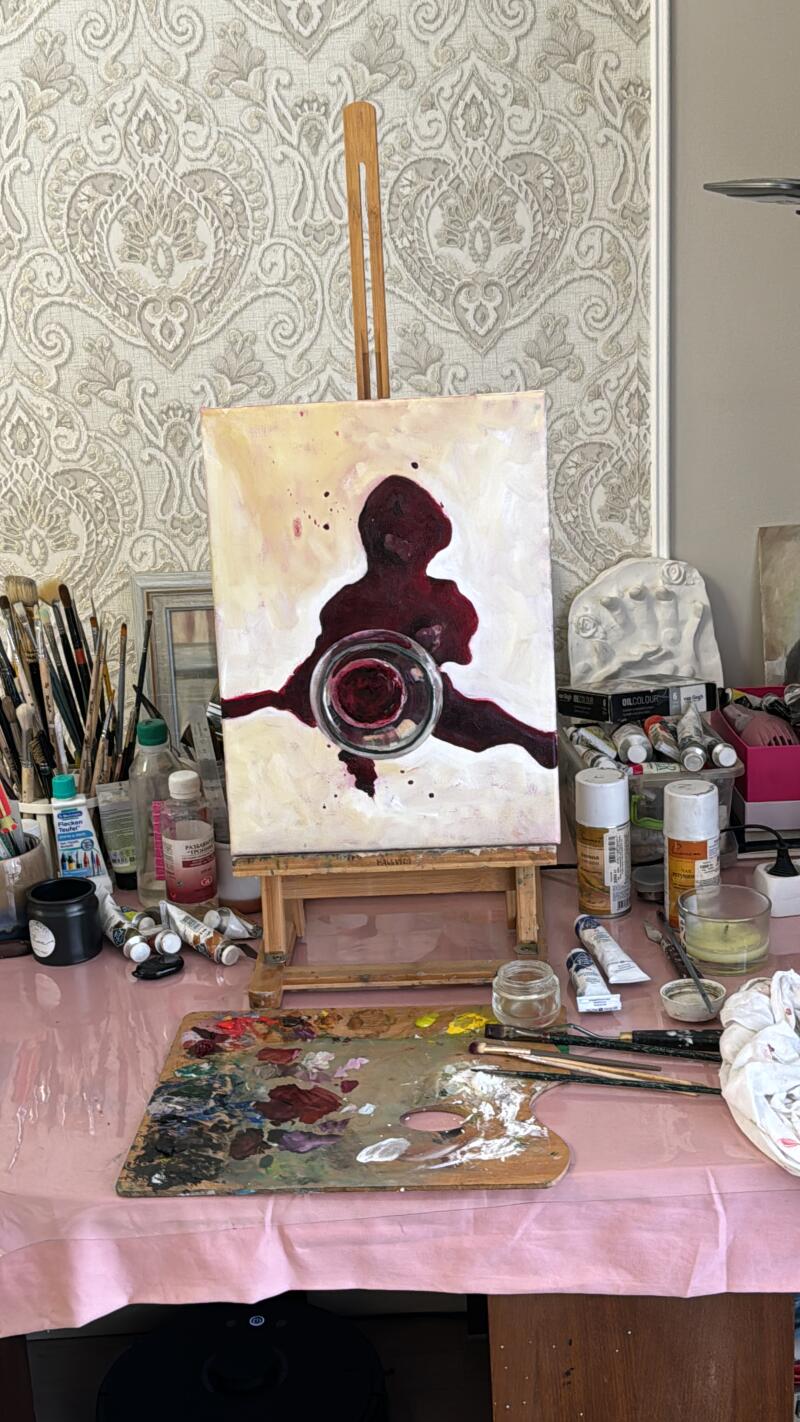 The image shows an easel with a canvas on which a silhouette of a person is depicted in burgundy color, with a glass object over the chest area, surrounded by various art supplies such as paints, brushes, and a palette with mixed colors on a pink-covered table.