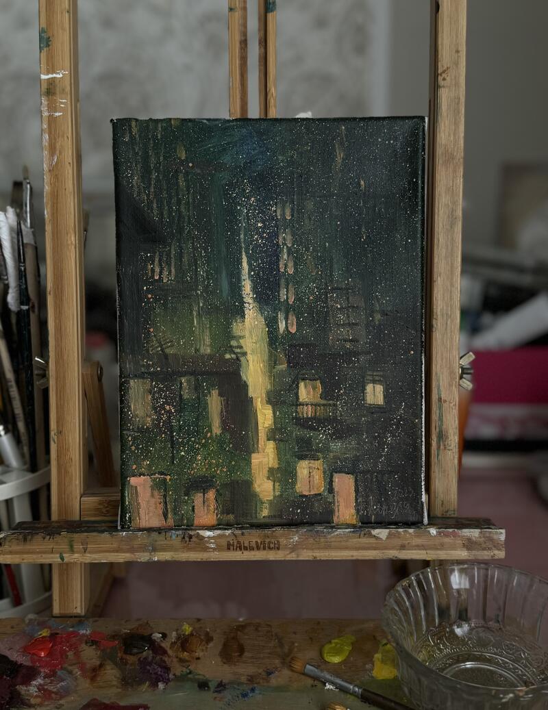 A painting of a cityscape at night sits on a wooden easel, with a palette of paints and a water bowl in the foreground. The painting depicts dark buildings illuminated by yellow lights, with the name 'HALEVICH' visible on the easel.
