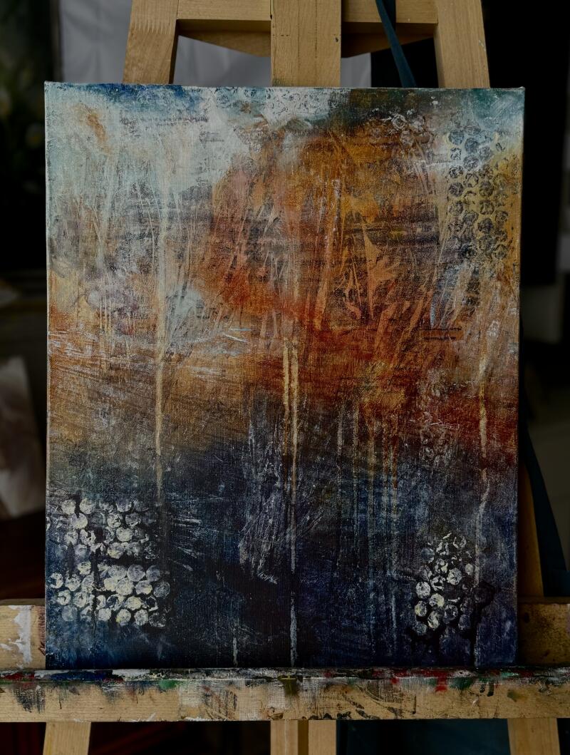 The painting on canvas features a blend of colors including white, blue, orange, and brown, with textural elements and dot patterns. It is displayed on a wooden easel.