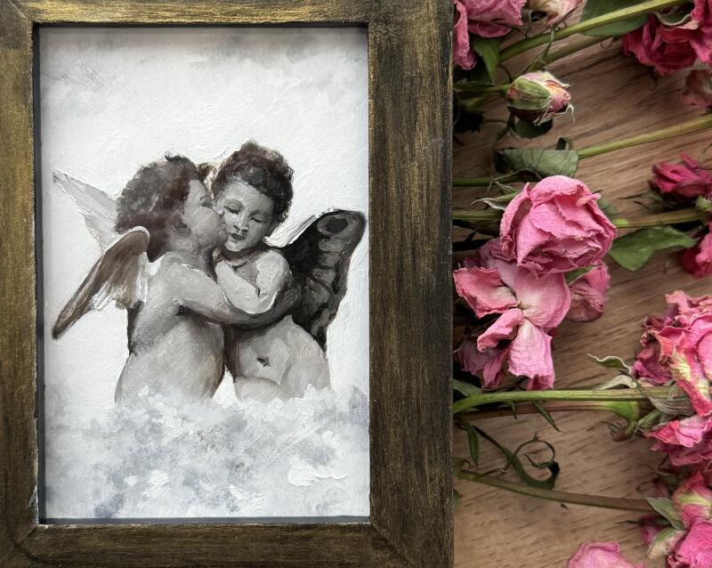 The image shows a framed painting of two angels embracing amidst clouds, with wilted pink roses scattered around the frame on a wooden surface.